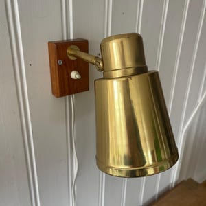 Könnte beinhalten: Eine Vintage-Wandleuchte mit Messingschirm und Holzsockel. Die Lampe hat einen konischen Schirm und einen kleinen Ein-/Ausschalter. Der Holzsockel ist rechteckig und hat einen warmen, rötlich-braunen Ton. Ein weißes Kabel erstreckt sich von der Lampe.