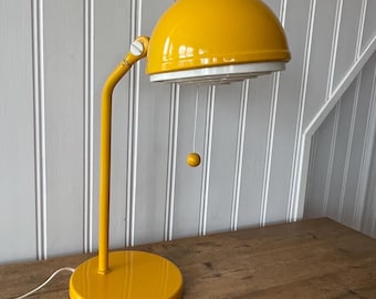 May include: A yellow table lamp with a round, dome-shaped shade and a white interior. The lamp has a white cord and a small, yellow ball hanging from the shade.