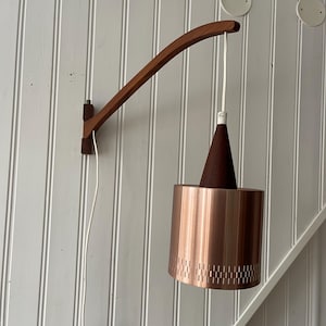 Può includere: Una lampada da parete in rame e legno con un braccio in legno curvo e un paralume in rame con un motivo perforato.