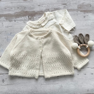 Op de afbeelding: Een crèmekleurige gebreide cardigan met een knoopsluiting en een delicaat kantpatroon. De cardigan ligt plat op een houten oppervlak. De tekst "Little French Knits" is zichtbaar in de afbeelding.