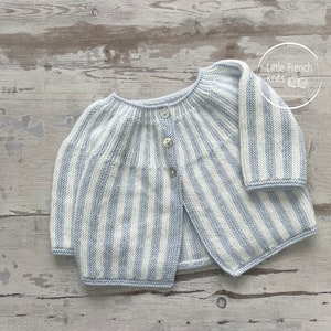 Op de afbeelding: Een gebreide baby-trui met lichtblauwe en witte strepen en knoopsluiting. De trui ligt plat op een houten oppervlak. De tekst "Little French Knits" is zichtbaar in de rechterbovenhoek van de afbeelding.
