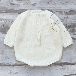 May include: A cream-coloured knitted baby romper with long sleeves and a button-up front. The romper has a round neckline. The garment is made of a soft, textured yarn and is displayed on a light-coloured wooden surface.