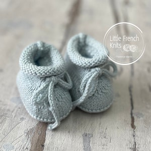 Könnte beinhalten: Ein Paar hellblaue gestrickte Babyschuhe mit einer Schleife an jedem Schuh. Die Schuhe liegen auf einer weißen Holzoberfläche.