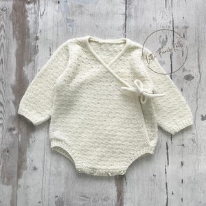 Peut inclure: Barboteuse pour bébé de couleur crème, avec des manches longues et un devant croisé, noué par un nœud. Le tissu tricoté présente un motif texturé. Le vêtement est présenté sur une surface en bois clair. Le texte "Little French Knits" est dans un cercle.