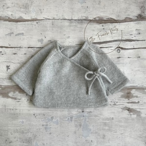 Może przedstawiać: Jasnoszary dzianinowy sweterek dla dziecka w stylu kopertowym z krótkimi rękawami i wiązaniem. Sweter wykonany jest z miękkiego, teksturowanego materiału i prezentowany na zwietrzałej drewnianej powierzchni. Tekst "Little French Knits" znajduje się w okręgu.