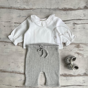 Könnte beinhalten: Ein Baby-Outfit mit einem weißen Langarmshirt mit Rüschenkragen und -bündchen, einer grauen Strickhose mit Kordelzug und passenden grauen Strickschuhen. Das Set wird auf einer hellen Holzoberfläche präsentiert.