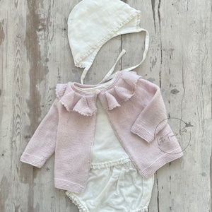 Può includere: Un completo per neonati composto da un cardigan rosa chiaro con colletto a balze, un body bianco con bordi in pizzo e un cappellino bianco abbinato. Il set è realizzato in morbido tessuto a maglia. Il tema generale è l'abbigliamento per bambini.