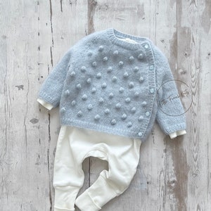 Puede incluir: Un conjunto de bebé de punto azul claro con diseño de pompón y pantalón blanco. La rebeca tiene cierre de botones y mangas largas. El texto "Little French Knits" es visible.