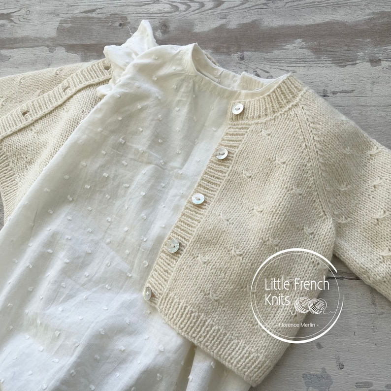 Puede incluir: Un c&aacute;rdigan de punto color crema con cierre de botones y un patr&oacute;n sutil. El c&aacute;rdigan est&aacute; superpuesto a una blusa de algod&oacute;n blanca con peque&ntilde;os puntos blancos.