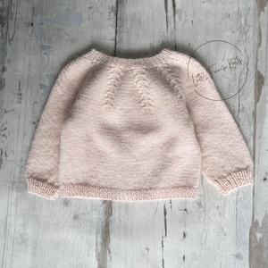 Peut inclure: Un pull rose clair pour bébé, tricoté, à manches longues. Le pull a une encolure ronde et un motif texturé sur l'empiècement. Les manches et l'ourlet ont une finition côtelée. Le texte "Little French Knits" est dans un cercle à droite.
