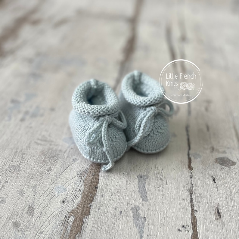 May include: A pair of light blue knitted baby booties with a tie at the top. The booties are on a light wood surface.