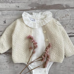 Op de afbeelding: Crèmekleurig babyvestje met een textuurgebreid patroon, gecombineerd met een witte bodysuit en een gerimpelde kraag. De outfit is geaccentueerd met een takje roze bloemen. De tekst "Little French Knits" is zichtbaar.
