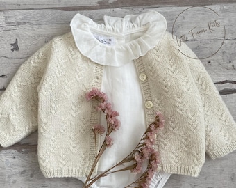 Pode incluir: Cardigã de bebê creme com um padrão de malha texturizada, combinado com um body branco e uma gola com babados. A roupa é realçada com um ramo de flores cor-de-rosa. O texto "Little French Knits" é visível.