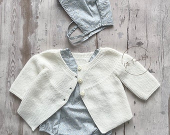 May include: A white knitted cardigan with a button closure, a light blue floral baby onesie, and a light blue floral bonnet. The outfit is laid out on a light wood background.