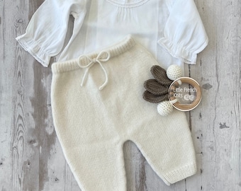 Op de afbeelding: Een wit gebreid babytopje met een kanten kraag en een crèmekleurige gebreide babybroek met een trekkoord. De broek ligt op een houten oppervlak met een klein houten speelgoed.