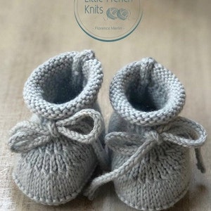 May include: A pair of gray knitted baby booties with a bow on each. The booties are tied together with a gray ribbon. The booties are on a wooden surface. The text "Little French Knits - Florence Merlin -" is visible in the background.