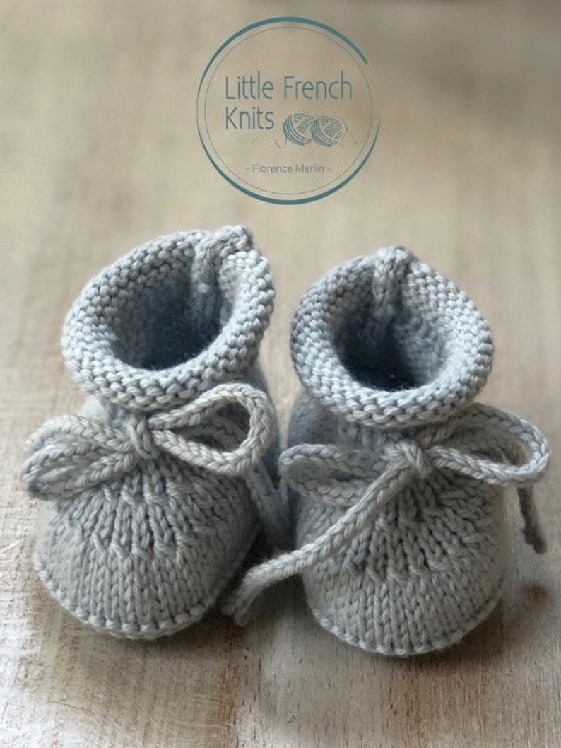 May include: A pair of gray knitted baby booties with a bow on each. The booties are tied together with a gray ribbon. The booties are on a wooden surface. The text "Little French Knits - Florence Merlin -" is visible in the background.