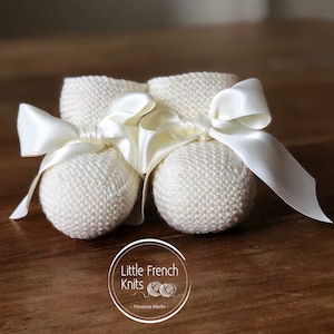Peut inclure: Une paire de chaussons en tricot blanc pour bébé avec des nœuds en ruban satiné. Les chaussons sont sur une surface en bois. Le texte "Little French Knits - Florence Merlin" est visible sur l'image.