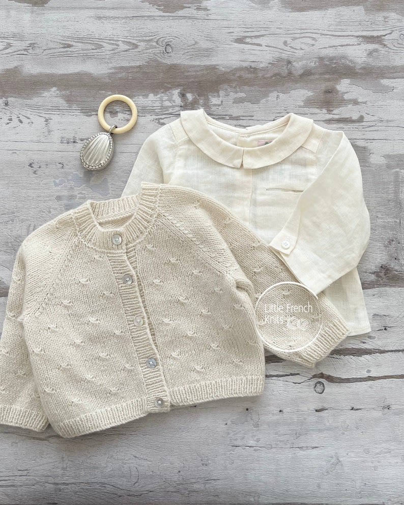 Puede incluir: Un c&aacute;rdigan de punto color crema con cierre de botones y una camisa de lino color crema con cuello Peter Pan. El c&aacute;rdigan tiene un patr&oacute;n texturizado y la camisa tiene un bolsillo en el lado izquierdo. Los art&iacute;culos est&aacute;n dispuestos sobre una superficie de madera clara.