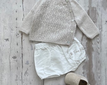 Pode incluir: Um conjunto de roupa de bebé bege claro, incluindo um cardigan de malha com botões, calças brancas e sapatos de bebé de camurça bege. O cardigan tem um padrão texturizado. O conjunto é exibido numa superfície de madeira clara.