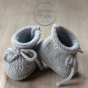 May include: A pair of gray knitted baby booties with a tied bow on the front. The booties are made by Little French Knits - Florence Merlin.