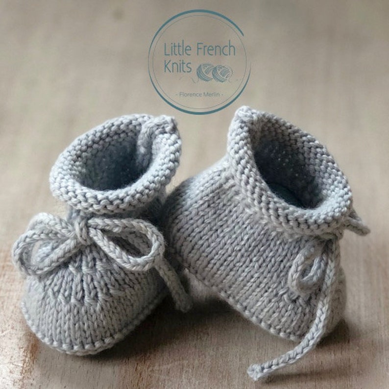 May include: A pair of gray knitted baby booties with a tied bow on the front. The booties are made by Little French Knits - Florence Merlin.