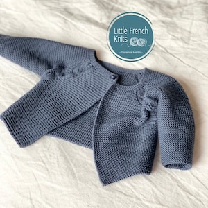 May include: A blue knitted baby cardigan with a button fastening and a cable knit design on the sleeves. The cardigan is laid flat on a white surface. The text "Little French Knits - Florence Merlin -" is visible in the top right corner of the image.