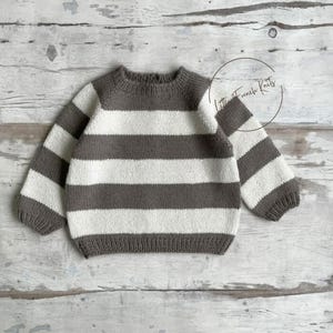 Peut inclure: Un pull en tricot à rayures grises et blanches. Le pull présente des rayures horizontales et des manches longues. Le texte "Little French Knits" est dans un cercle en haut à droite. Le pull est présenté sur une surface en bois clair.