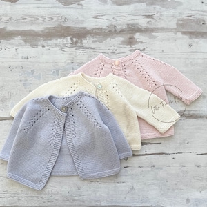 Peut inclure: Trois cardigans en tricot pour bébé dans différentes couleurs : violet clair, crème et rose clair. Chaque cardigan a une fermeture à boutons et un motif ajouré délicat.
