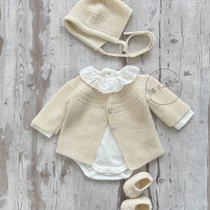 May include: A cream knitted baby cardigan with a white bodysuit, a cream knitted hat with a tie, and cream knitted booties. The outfit is laid out on a whitewashed wood background.