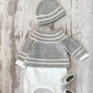 May include: A grey knitted baby sweater with white stripes, a matching grey knitted hat with white trim, and white knitted booties. The outfit is laid out on a light wood background.