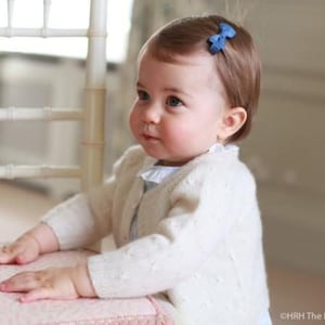 Puede incluir: Una ni&ntilde;a peque&ntilde;a con cabello casta&ntilde;o claro lleva un c&aacute;rdigan blanco con un lazo azul en el pelo. Est&aacute; de pie junto a una silla blanca con un coj&iacute;n rosa.