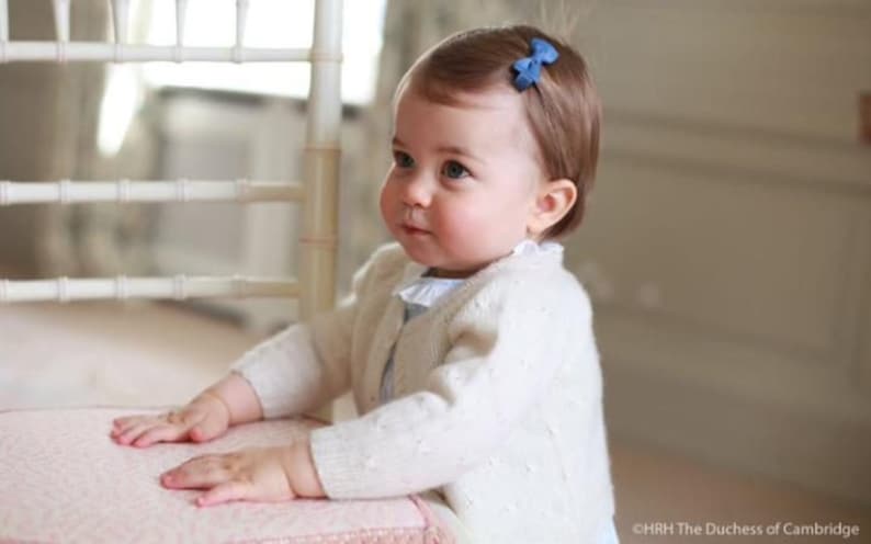 Puede incluir: Una ni&ntilde;a peque&ntilde;a con cabello casta&ntilde;o claro lleva un c&aacute;rdigan blanco con un lazo azul en el pelo. Est&aacute; de pie junto a una silla blanca con un coj&iacute;n rosa.