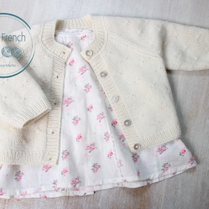 Puede incluir: Un c&aacute;rdigan de punto color crema con botones de perla y un delicado ribete de encaje alrededor del escote. El c&aacute;rdigan est&aacute; colocado sobre una superficie de madera con un vestido de estampado floral debajo.