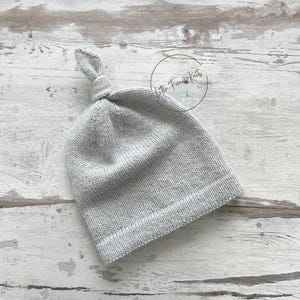 May include: A light gray knitted baby hat with a knotted top. The hat is made of soft yarn and has a ribbed brim. The hat is displayed on a light-colored wooden surface. The text "Little French Knits" is in a circle in the background.