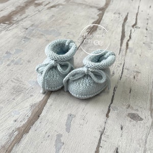May include: A pair of light blue knitted baby booties with a bow on each. The booties are on a light wood background. The text "Little French Knits" is visible in the top right corner of the image.