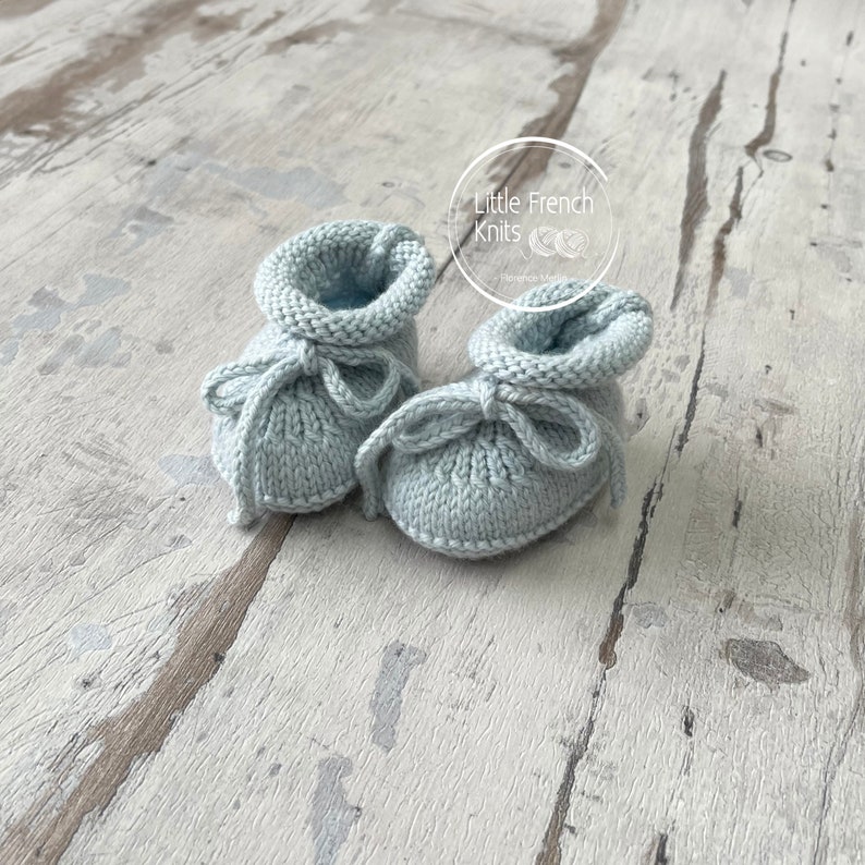 May include: A pair of light blue knitted baby booties with a bow on each. The booties are on a light wood background. The text "Little French Knits" is visible in the top right corner of the image.