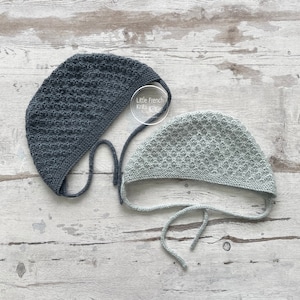 Peut inclure: Deux bonnets de bébé tricotés, l'un en gris foncé et l'autre en bleu clair, avec des liens. Les bonnets sont sur une surface en bois blanc. Le texte "Little French Knits" est visible sur l'image.