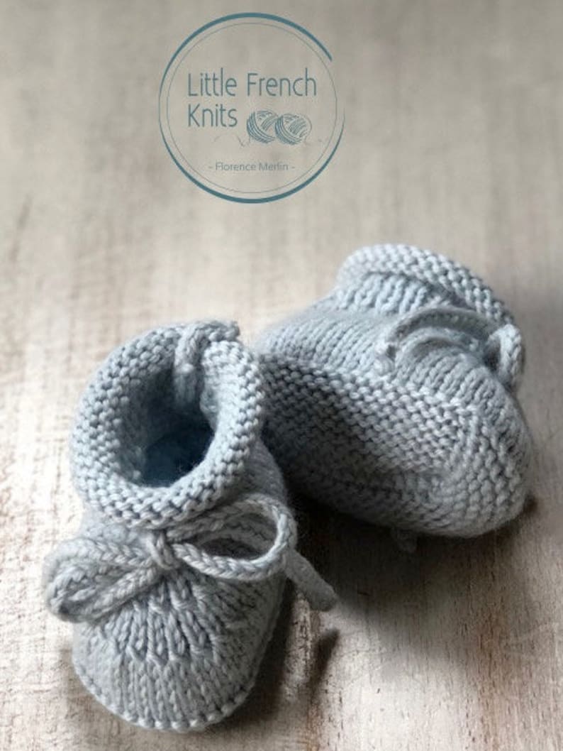 May include: A pair of gray knitted baby booties with a bow tie detail. The booties are on a wooden surface.  Little French Knits - Florence Merlin -