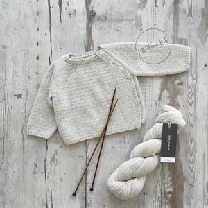 May include: A light grey knitted baby sweater with a single button closure on the shoulder. The sweater is laid out on a light wood surface with knitting needles and a ball of yarn.