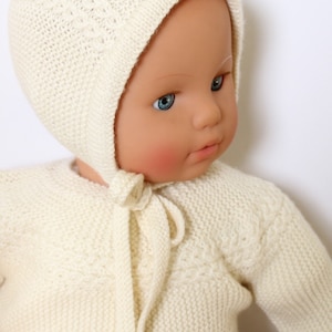 Op de afbeelding: Een crèmekleurige gebreide baby-poppenoutfit, inclusief een muts en een trui. De muts wordt onder de kin vastgebonden met een bijpassende gebreide stropdas. De trui heeft een ronde halslijn en lange mouwen. De pop heeft blauwe ogen en roze wangen.