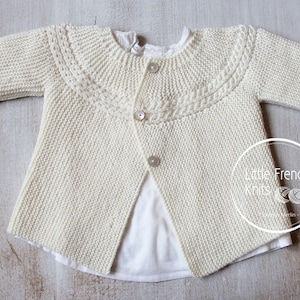 May include: A cream knitted cardigan with a button fastening and a decorative cable knit pattern around the neckline. The cardigan has long sleeves and a slightly cropped length.