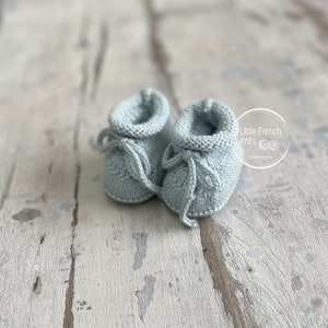 May include: A pair of light blue knitted baby booties with a tied bow on each. The booties are on a light wood surface.