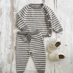 Könnte beinhalten: Ein gestreiftes Strick-Set aus braunem und weißem Pullover und Hose mit einem Knopfverschluss am Pullover. Das Set liegt auf einer weißen Holzoberfläche mit einem Paar beigen Schuhen.