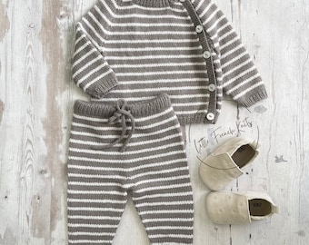 May include: A striped knit sweater and pants set in brown and white with a button closure on the sweater. The set is laid out on a white wooden surface with a pair of beige shoes.