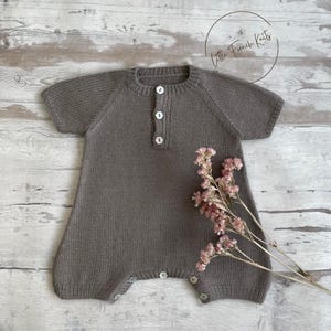May include: A knitted grey baby romper with short sleeves and white buttons down the front. The romper has buttons at the bottom for easy nappy changes. The words "Little French Knits" are in a circle in the upper right corner. A sprig of pink flowers is on the right.