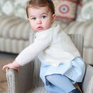 Puede incluir: Una ni&ntilde;a peque&ntilde;a con un c&aacute;rdigan de punto blanco y un vestido azul claro con estampado floral se sienta en una silla de mimbre. La ni&ntilde;a lleva medias azules y zapatos azul oscuro con una raya blanca. La ni&ntilde;a tiene el pelo rojo y mira por encima del hombro.