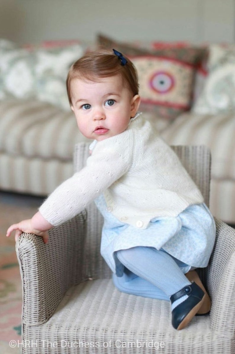 Puede incluir: Una ni&ntilde;a peque&ntilde;a con un c&aacute;rdigan de punto blanco y un vestido azul claro con estampado floral se sienta en una silla de mimbre. La ni&ntilde;a lleva medias azules y zapatos azul oscuro con una raya blanca. La ni&ntilde;a tiene el pelo rojo y mira por encima del hombro.
