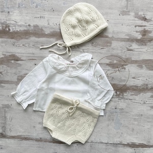 May include: A three-piece baby outfit set in cream. The set includes a knitted bonnet with ties, a long-sleeved white blouse with a ruffled collar, and matching knitted shorts. The shorts have a drawstring waist. The words "Little French Knits" are in the image.