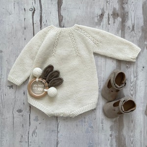 Op de afbeelding: Een crèmekleurige gebreide baby romper met een kabelbreipatroon. De romper ligt plat op een licht houten oppervlak. Naast de romper ligt een houten baby rammelaar met bruine en witte pompoenen. De rammelaar heeft de tekst "Little French Knits" erop. Twee bruine leren babyschoentjes liggen aan de rechterkant van de afbeelding.